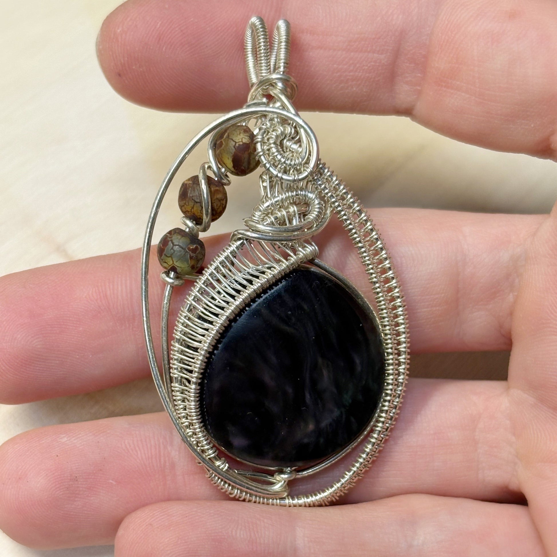 Hand holding a silver wire-wrapped pendant with a dark stone and smaller beads.