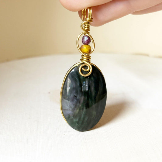 Hand holding a pendant with a large green stone and smaller colored glass beads on top, against a neutral background.-p