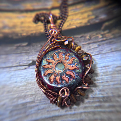 A close-up of a handmade copper wire pendant necklace featuring a sunburst design over a dark, iridescent circular background, resting on a rustic weathered wood surface.