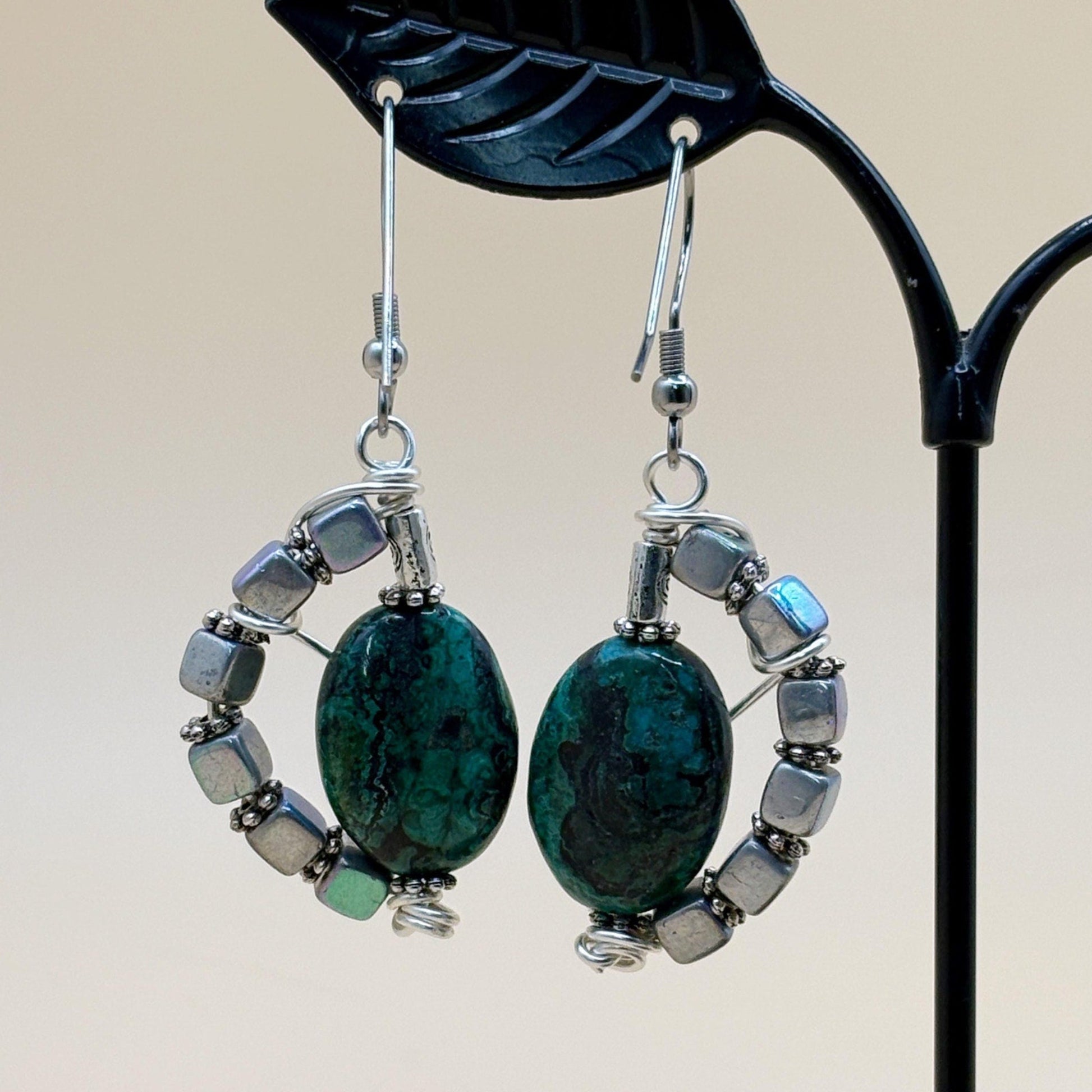 Pair of earrings with green stones and silver accents on a stand.