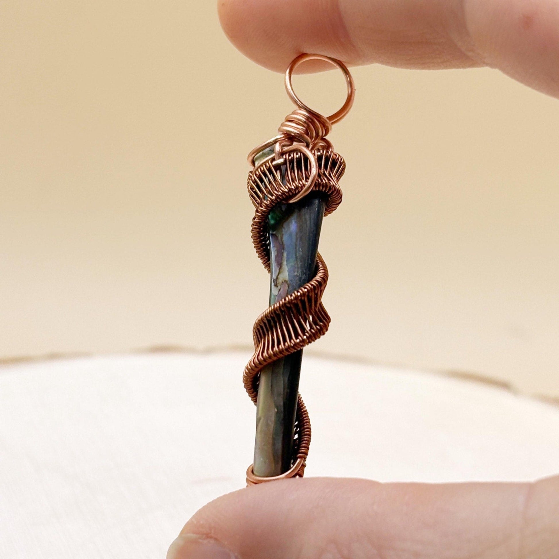 Copper-wrapped pendant with a central shell held between fingers against a neutral background