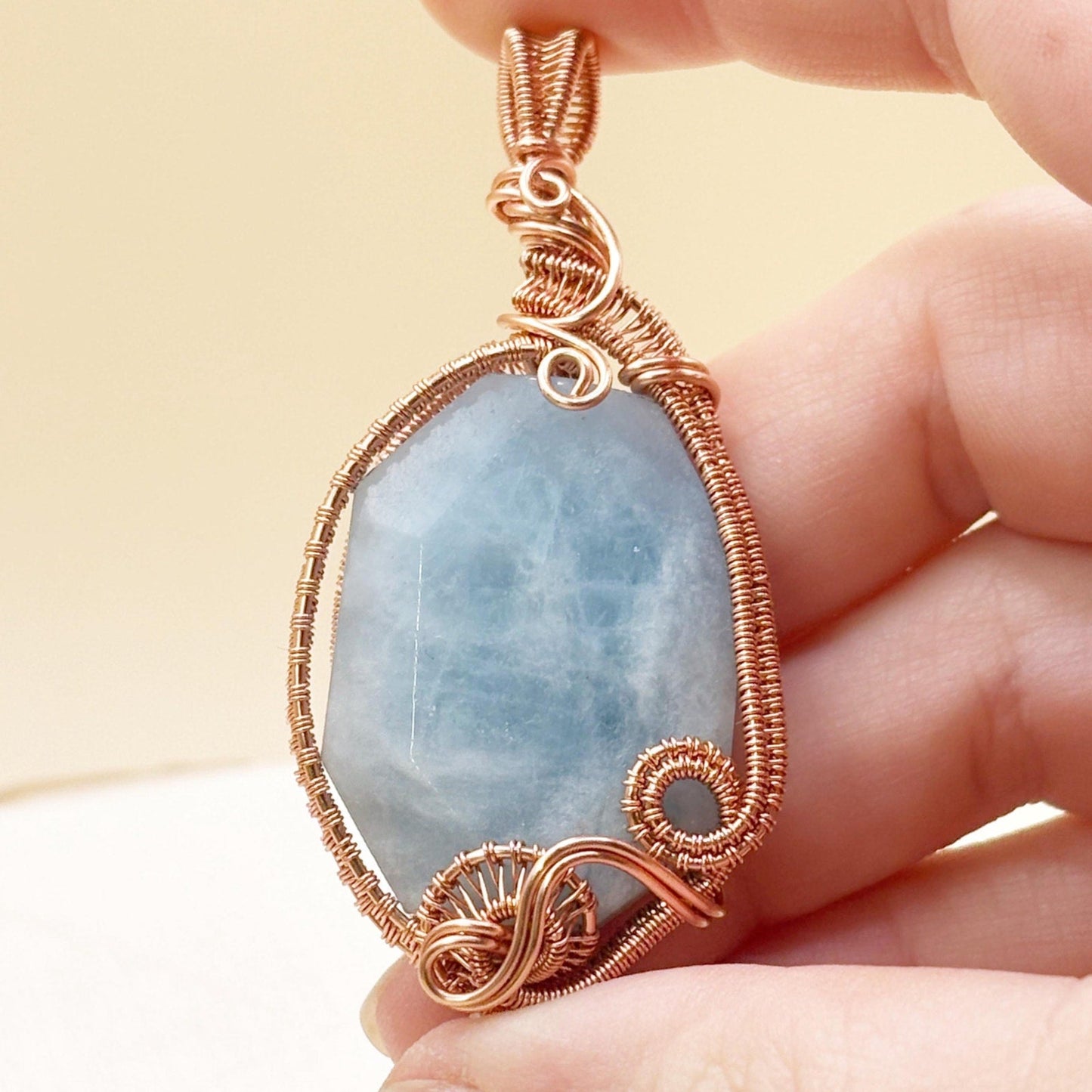 Hand holding a pendant with a blue stone and gold wire wrapping against a beige background