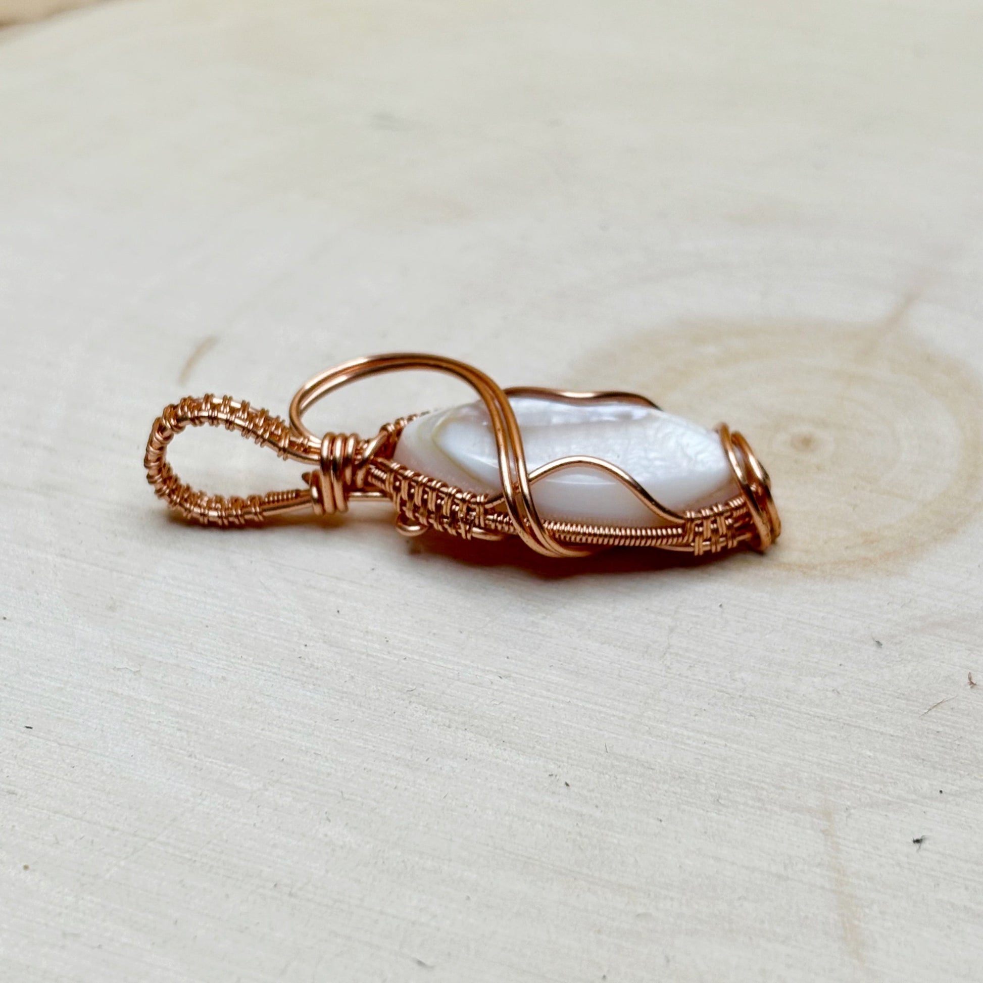 Necklace pendant with a mother of pearl shell wire wrapped in shiny copper on a light wooden background