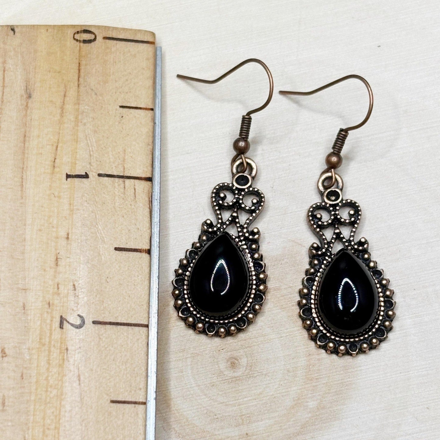 Pair of black teardrop earrings with bronze detailing on a wooden surface with a ruler for scale.