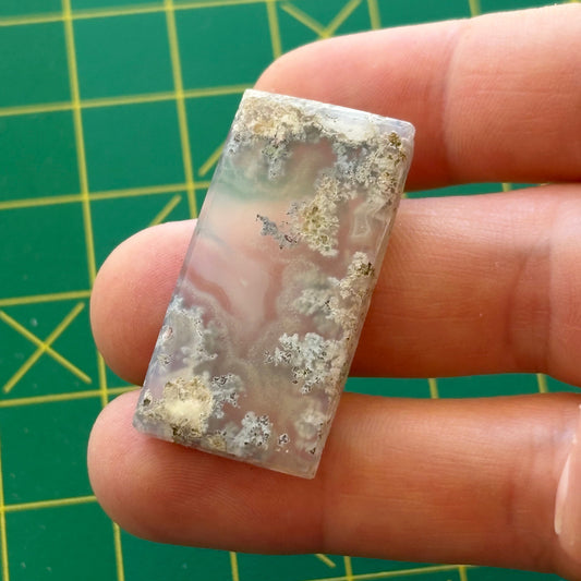 Rectangular crystal stone held between fingers against a green grid background-p
